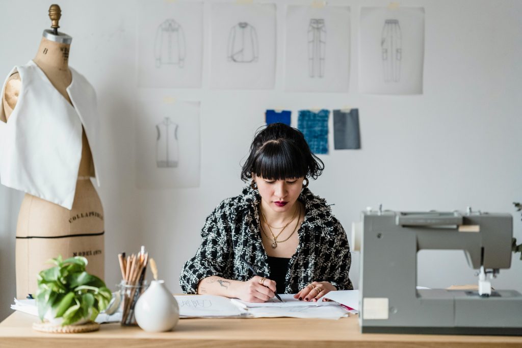 Imagem de uma jovem desenhando um em um ateliê. O ambiente conta com um manequim, uma máquina de costura, materiais de desenho, além de croquis e amostras de tecido pregadas na parede.