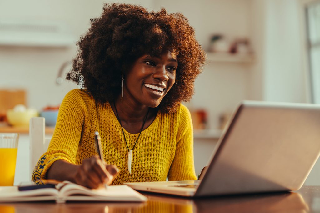 mulher negra sorri olhando para o notebook enquanto faz anotações em um caderno