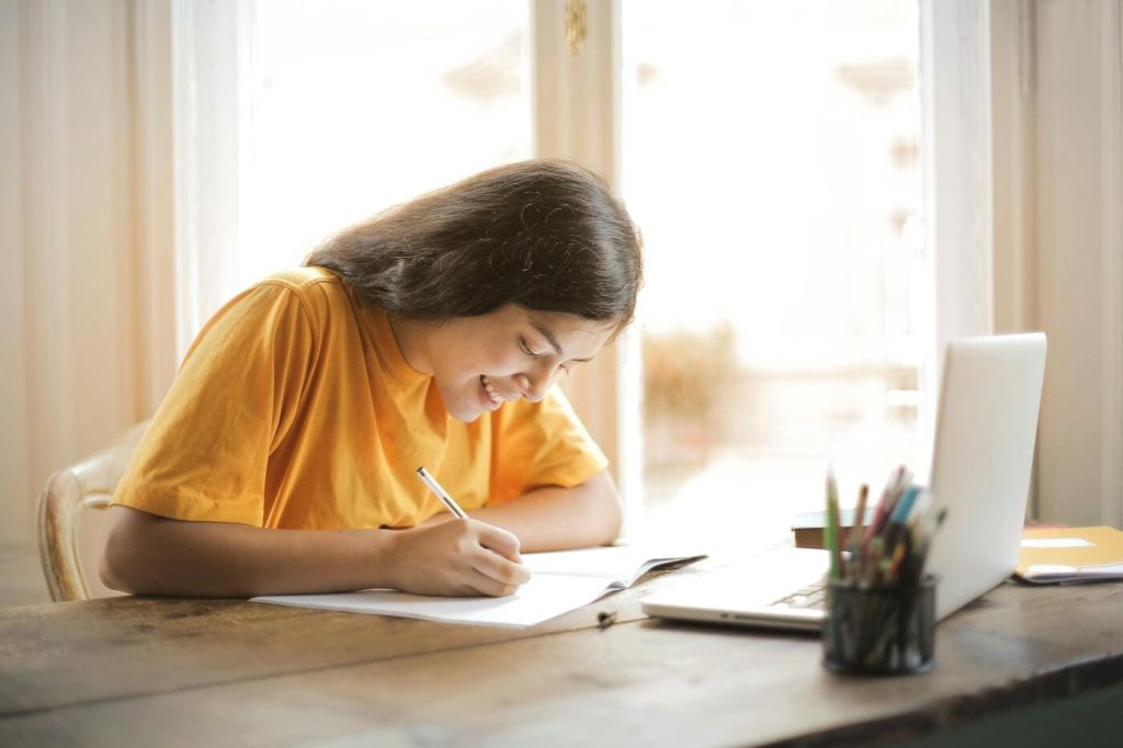 Mulher jovem escolhendo uma faculdade no UniSENAI. Ela está escrevendo em um caderno e sorrindo. Sobre a mesa, há um notebook e um porta canetas.